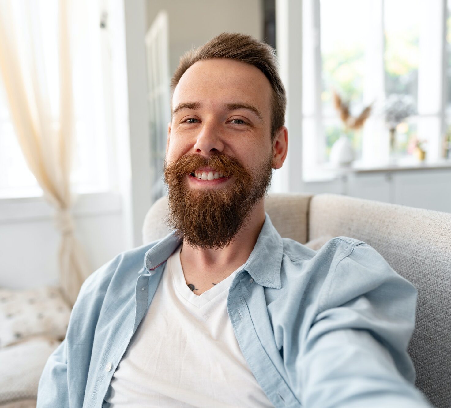 Bearded happy young man smiling and taking selfie photo while sitting on sofa at home.